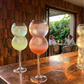 Three bubble-shaped cocktail glasses with colorful drinks on a wooden table, with people and greenery in the background.