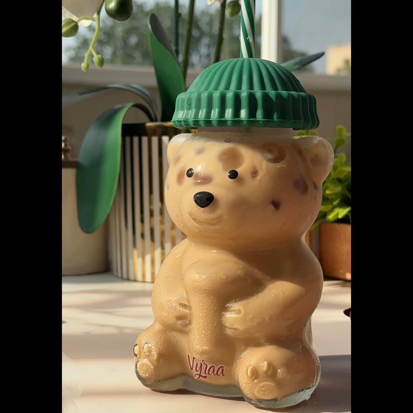 A beige bear-shaped glass bottle with a green ribbed lid and a green-and-white striped straw, set on a light surface with a window and plants in the background.