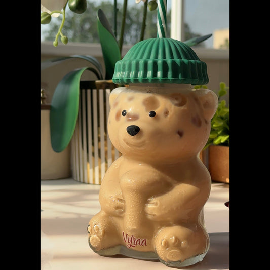 A beige bear-shaped glass bottle with a green ribbed lid and a green-and-white striped straw, set on a light surface with a window and plants in the background.