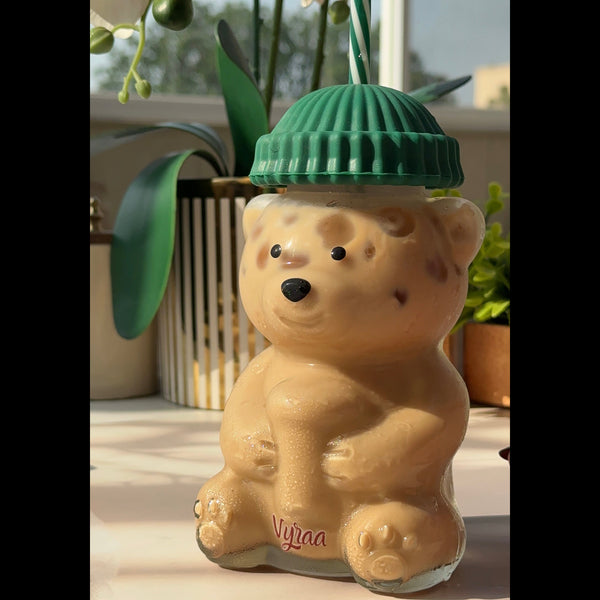 A beige bear-shaped glass bottle with a green ribbed lid and a green-and-white striped straw, set on a light surface with a window and plants in the background.