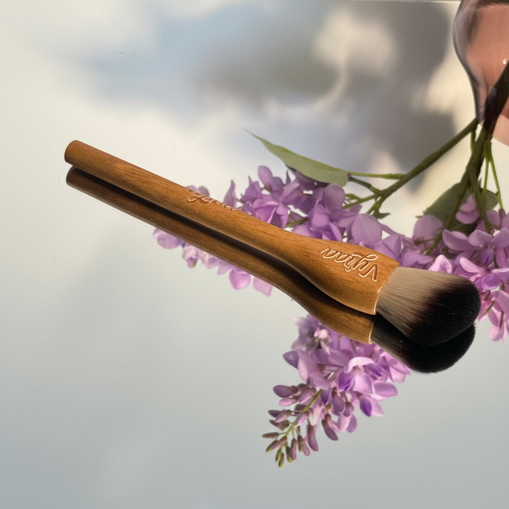 Makeup brush with wooden handle and pink bristles on a reflective surface with flowers.