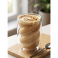 Glass mug with a spiral of coffee on a wooden coaster with a spoon, plant, and window in the background.