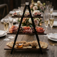 Three-tiered wooden appetizer tray with cured meats, cheeses, and crackers on a wooden table.